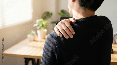 Man holding painful shoulder while sitting at home office desk, muscle strain and workplace injury awareness concept