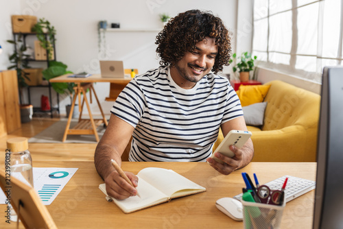 Young adult Indian man student studying with smartphone and notebook at home desk. Focused learner combining mobile technology and online education resources. Education and e-learning concept.