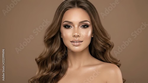 Beautiful woman with wavy brown hair looking at camera. Studio portrait with neutral brown background, focusing on professional makeup and glowing skin. Concept of beauty salon, cosmetics, skincare.