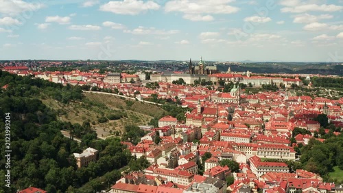 Prague Castle complex and Saint Vitus Cathedral rise above historic red tile roofs in Mala Strana district. European capital city panorama under sunny blue sky creates vibrant travel background.