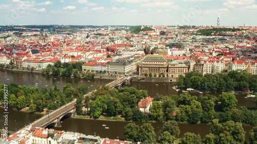 Prague cityscape featuring Vltava river, National Theater and historic bridge. Red tile roofs fill urban panorama under sunny blue sky. European capital city travel destination background.