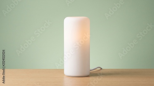 A modern minimalist cylindrical white desk lamp emitting a warm, soft glow, placed on a light wooden table against a subtle sage green background.