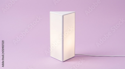 A minimalist white triangular prism table lamp glowing with warm light against a smooth pastel purple background. Perfect modern desk light or nightstand decor