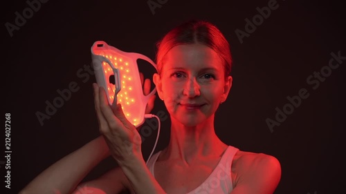 woman holding infrared light therapy LED mask then move it away and shows the face smiling.  relaxed smiling face looking at the camera, black  background. Contrast red light. horizontal composition v