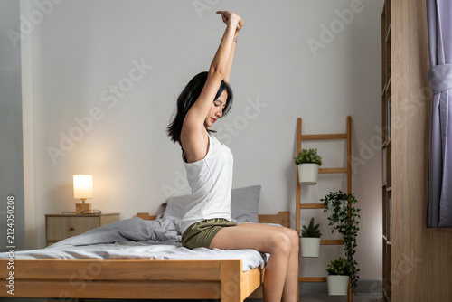 Fotografie Young Asian woman sitting on bed stretching arms morning wake up, happy female r