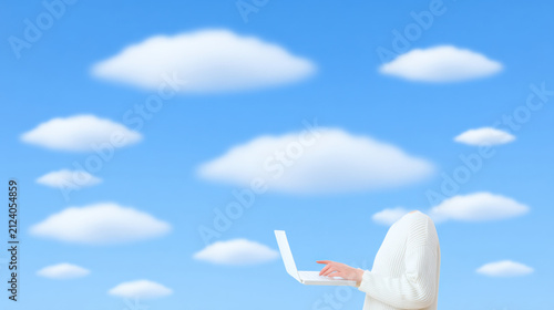 A person with no visible head works on a laptop against a bright blue sky filled with fluffy white clouds.