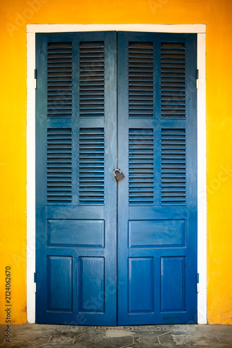 Closed vintage blue wooden louvered doors on yellow facade of ancient building in Hoi An Vietnam, Vietnamese traditional style exterior entrance with doors closed on metal lock against yellow wall.