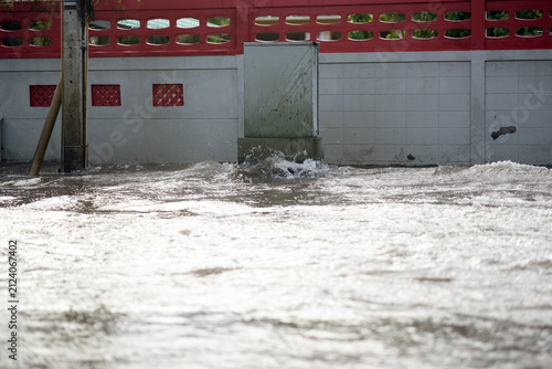 Rushing floodwaters washing along an urban street or canal next to a decorated concrete property wall during a heavy downpour or flood event.