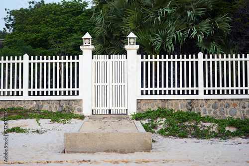 Charming white wooden picket fence and closed gate on a sandy beach path leading to lush green trees.