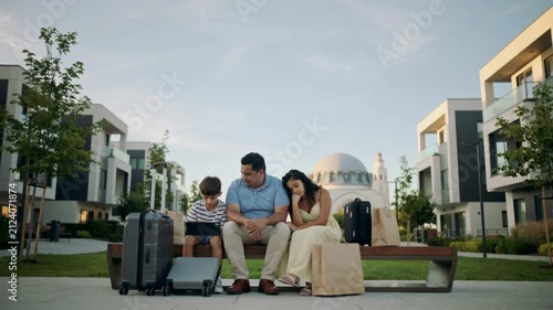 Middle Eastern family sitting on bench with suitcases and tablet outdoors with copy space eye level