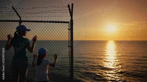 Mother and daughter standing by chain link fence overlooking ocean at sunset, static shot