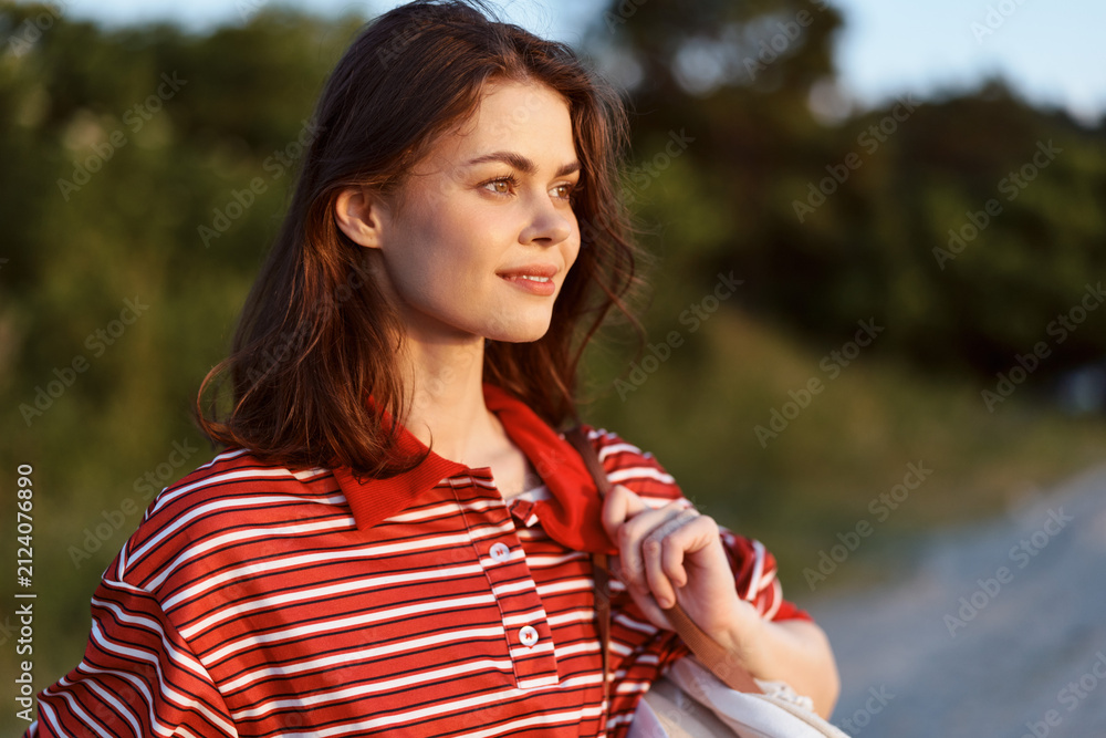 Obraz premium Young woman enjoying lifestyle outdoors at sunset. Positive girl wearing striped shirt standing in nature landscape. Warm sunlight illuminates her face during summer evening walk.