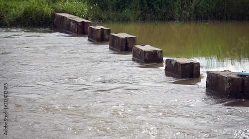 Muddy river water flowing rapidly through a small concrete dam barrier. Rural irrigation system and water management infrastructure footage showcasing stream current movement in countryside