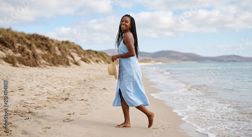 Portrait of a smiling young Black woman in a blue summer dress holding a straw hat on a sandy beach