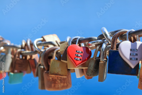 Fotografie Red heart shaped combination love lock attached to chain with other padlocks against blue sky