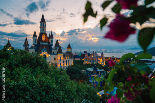 Illuminated castle-inspired of Vinpearl in VinWonders theme park overlook a scenic bay at dusk, creating a magical waterfront cityscape with mountains, dramatic clouds, and evening lights.