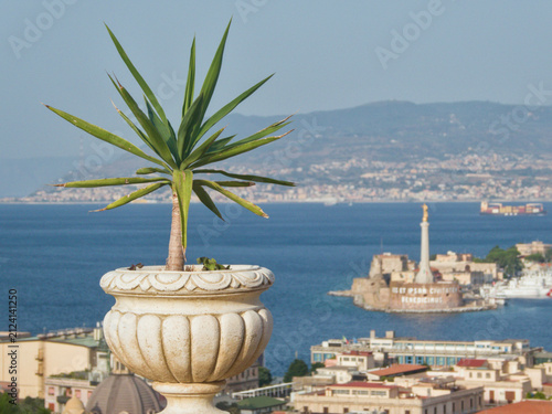 Canvastavla Potted green plant in decorative ornamental stone planter featuring scenic background view of Messina harbor and Madonnina monument in Sicily, Italy on sunny summer day