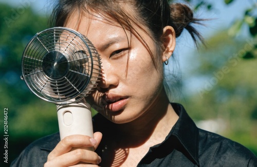 Woman holding portable fan outdoors. Young woman cooling herself with portable electric fan outdoors on sunny summer day, heatwave and personal cooling concept