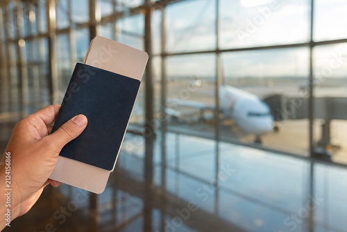 Airport Travel Essential: A hand holds a passport and boarding pass inside a modern airport terminal, with an airplane visible through large windows, ready for departure.