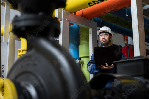 Focused professional male industrial engineer uses a laptop computer to inspect heavy machinery and colorful utility pipes during maintenance inside a modern manufacturing plant facility.