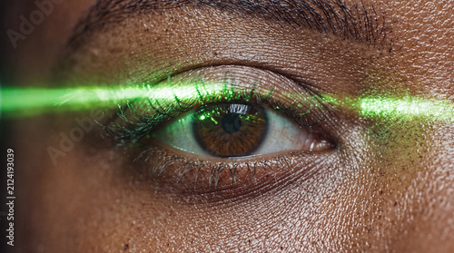 Extreme close-up of a brown eye with a bright green laser beam cutting horizontally across the iris, symbolizing technology and vision.
