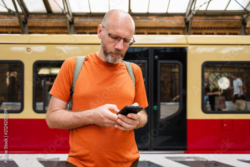 Man using smartphone at train station platform with yellow and red train