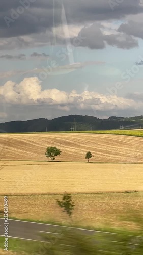 Vertical view through the window of a moving vehicle overlooking a summer countryside with green and yellow fields, scattered trees, rolling hills, and dramatic clouds.