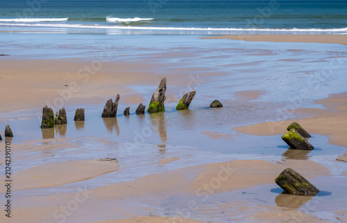 Remains of a wooden shipwreck exposed at low tide on Bamburgh Beach, Northumberland coast, weathered timbers revealed in shifting sand under coastal light