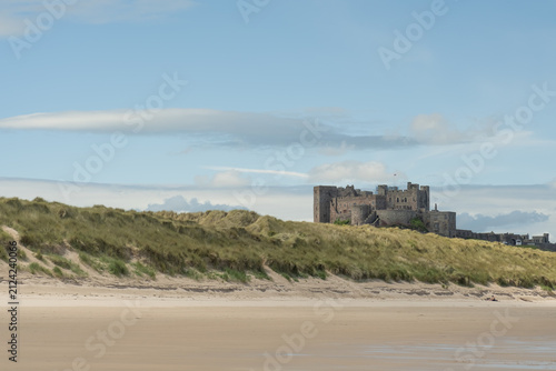 Historic medieval castle set among sand dunes on the North East coast, dramatic coastal setting with weathered stone architecture against shifting sands and sky.