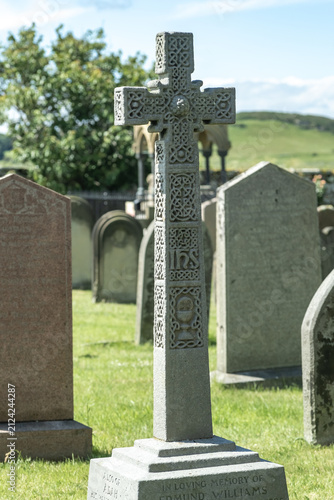 Close and selective focus on an ornate Celtic cross in a graveyard, with blurred bokeh background, highlighting intricate stone carving and quiet, atmospheric mood.