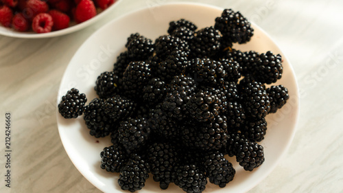 Fresh blackberries in a white bowl on a marble counter with raspberries blurred in the background.