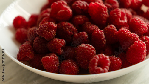 Fresh ripe raspberries piled high in a white ceramic bowl on a light marble tabletop, top down view.