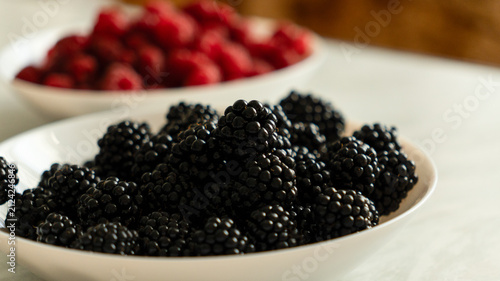 Fresh blackberries in a white bowl on a marble counter with raspberries blurred in the background.