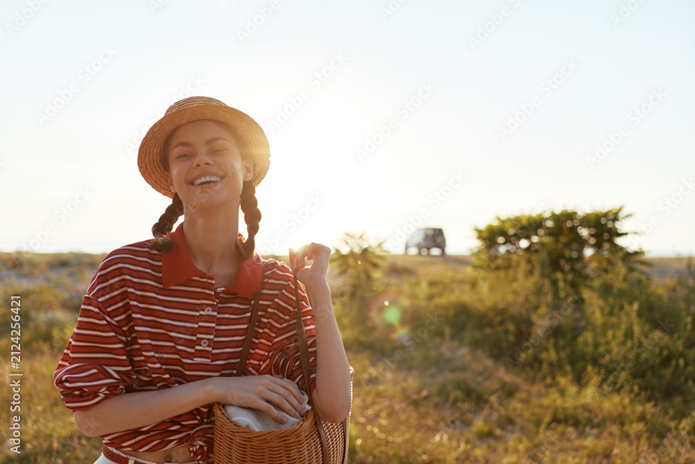 Obraz premium A joyful woman in a striped shirt and straw hat stands in a sunlit field with a woven basket, embracing carefree outdoor lifestyle and warm summer vibes. The scene radiates ease. Warm.