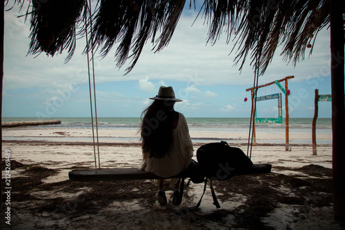 Fotografie Woman relaxing on a wooden swing under a thatched roof palapa looking at the ocean