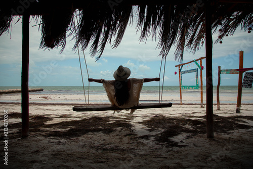 Принти на полотні Woman relaxing on a wooden swing under a thatched roof palapa looking at the ocean