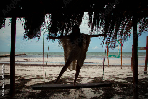 Bild auf Leinwand Back view of a woman standing on a beach swing with arms outstretched under a thatched shelter
