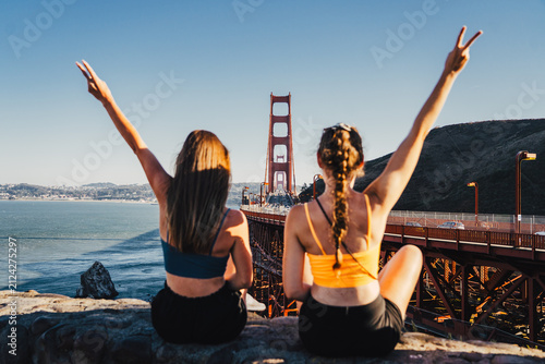 Foto Two young women tourists sitting on viewpoint ledge making victory peace sign wi