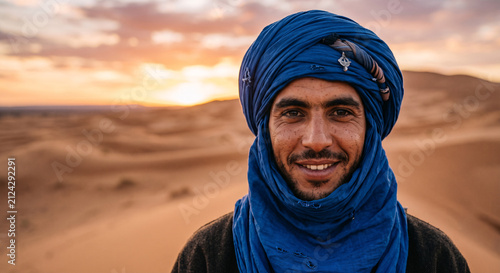 Portrait of smiling Tuareg man in blue traditional turban backgrounded by Sahara desert dunes at sunset