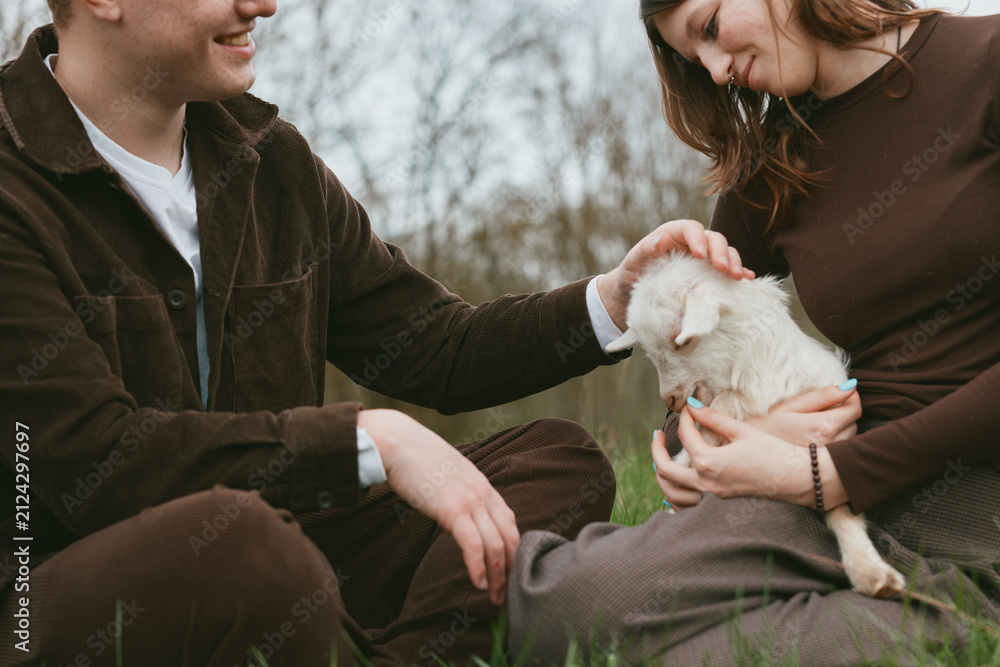 Obraz premium Couple outdoors with a goat kid, gentle interaction, warm atmosphere, lifestyle moment, close-up of care and joy, natural light and grassy backdrop evoke serenity and companionship.