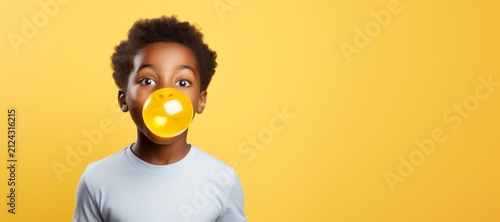 Young African American boy blowing a large yellow bubble with gum, wearing a light blue shirt, against a vibrant yellow background, capturing a playful and joyful moment of childhood