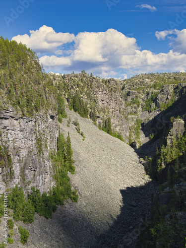View of the dramatic Jutulhogget canyon featuring steep rocky walls, scree slopes, and pine trees under a blue sky in Alvdal, Innlandet, Norway.