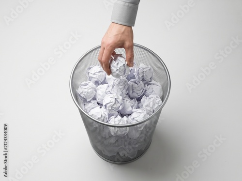 Hand throwing crumpled paper into office trash bin. Person discarding waste documents in metal recycling container for eco-friendly disposal.