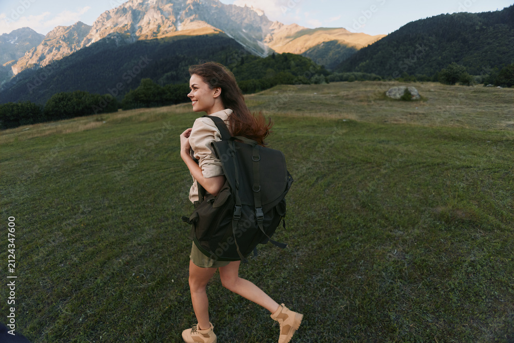 Obraz premium Cheerful woman outdoors, hiking with a backpack, grassy field and distant mountains in the background, sunlight on her hair, relaxed stride, natural colors and breeze