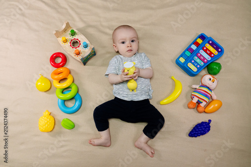 Cute 11 month baby boy lying among various playful colorful toys on beige blanket. Infant surrounded by vivid objects participating actively in sensory exploration and learning. 