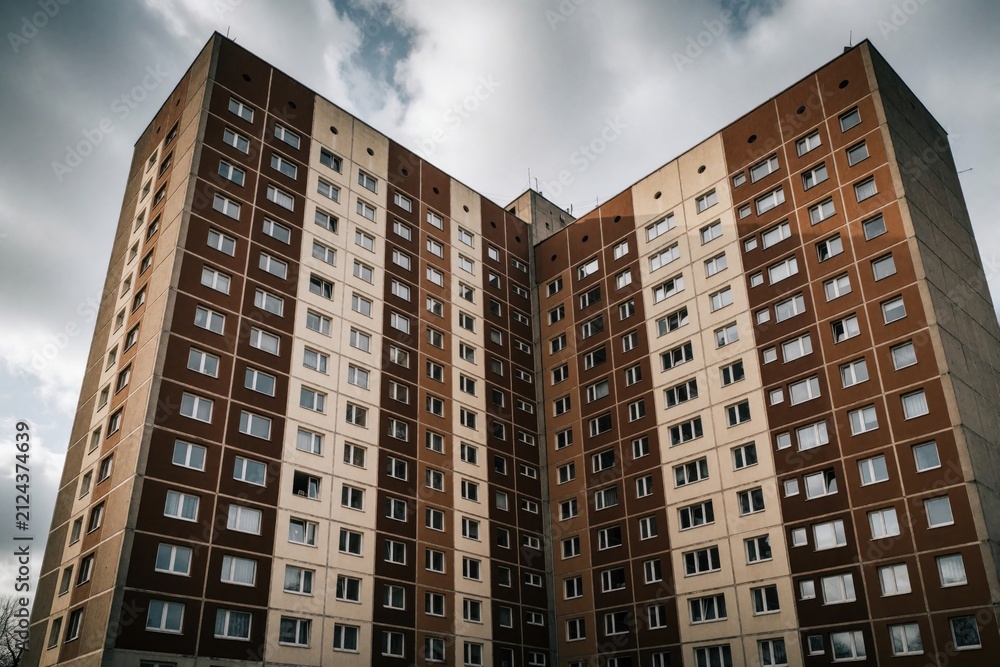 Fototapeta premium Tall concrete residential block with brown and beige panels towers over city. Many windows and balconies mark apartments in this old communist era building complex.
