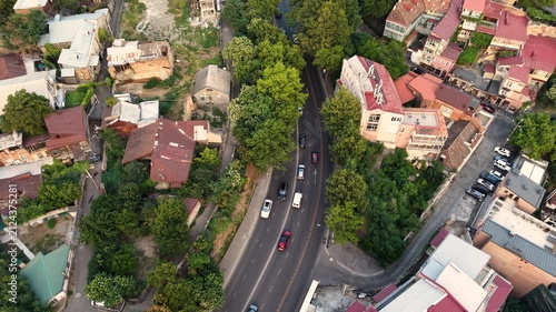 Top-down drone shot looking down at cars driving along a curved asphalt road cutting through an old city district filled with traditional tile-roof houses, green trees, and cozy narrow streets