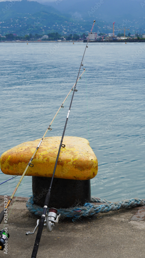 Obraz premium fishing rods resting on a yellow mooring bollard at a harbor pier overlooking the sea and distant mountains