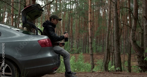 Man sitting on car trunk and drinking coffee from thermos in pine forest, audio included