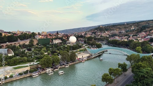 Breathtaking drone panorama over Tbilisi showcasing major travel attractions, including the modern glass bridge over the river, green park walkways, scenic hills, and historic city landmarks 